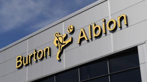 External view of the Pirelli Stadium with a Burton Albion logo shown fixed to the side of a stand.