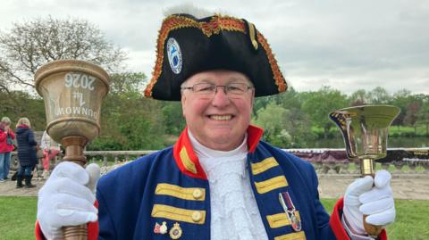 A man with a classic town crier's outfit with blue and red colours and a black hat. His white-gloved hands are ringing two bells.