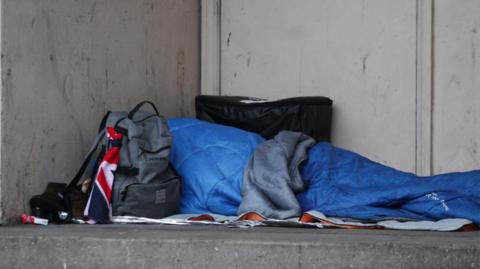 A homeless person in a blue sleeping bag lying outside on a concrete floor, with a backpack in front of their face.