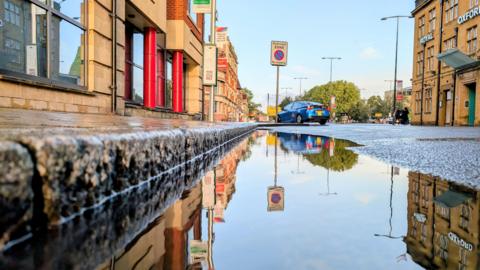 An Oxford street scene captured from a low angle just above a puddle. You can see a blue car and street signs and there are buildings on either side of the road. The camera is at kerb level and the scene is reflected in the water. 