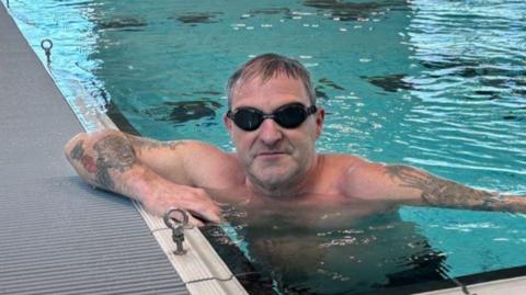 A man with dark hair and dark goggles is in the pool at one edge, with his arms up over the side