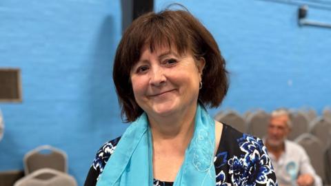 A woman wearing a blue scarf is standing in a sports hall with a blue wall. There are chairs and other people behind her. She is smiling for the photograph.