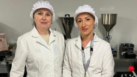 Employees Marzena and Dorota are pictured in factory PPE gear, a white protective long jacket and a white cap. They are standing together looking into the camera.