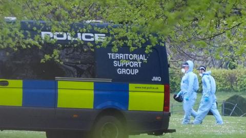 A dark blue police van and two men in blue forensics suits walking behind it in a park.