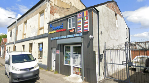 Google street view picture of the outside of SK First Stop. It's a small building, painted grey with a white door whilst the shop name is in red, blue and yellow writing above the door.