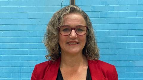 A woman with shoulder length, curly blonde hair, stands in front of a blue-painted brick wall. She wears a red jacket over a black top and has red-rimmed glasses.