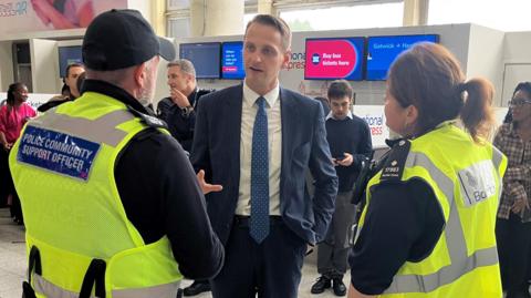 Mike Tapp is standing in an airport in a navy suit with a blue tie, which has white dots on it. He is talking to two people in yellow high-vis vests. One vest says "police community support officer" on the back and the other says "Border Force".