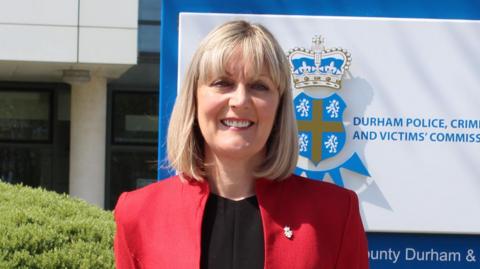 Head and shoulder shot of a smiling woman with a blonde bob and wearing a red jacket over a black top. Behind her is a Durham Police sign.