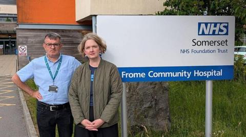 A man and a woman standing side by side. The man has his hands on his hips and the woman has her hands in front of her. They are standing in front of the sign for Frome Community Hospital.