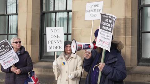 Three people, one man and two women, all carrying placards protesting council tax rises. The man also holds a megaphone and is speaking into it.