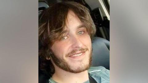 Sam Rimmer has long, light brown hair and a short beard. He has green/blue eyes and smiles for the  camera in what appears to be a selfie taken while in a vehicle.