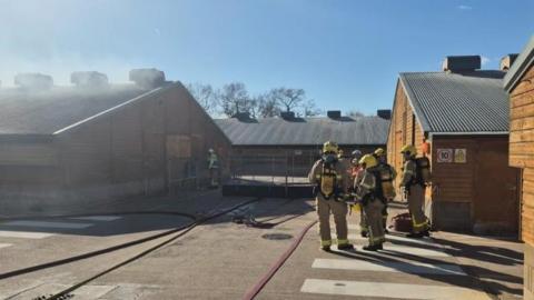 A group of firefighters in brown uniforms and carrying equipment. They are outside a number of barns, one which has smoke coming out of it. 