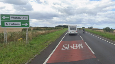 A single carriageway stretch of the A1 has a sign to Wandylaw pointing to the left and a sign to Newham to the right. A caravan is ahead with two motorcyclists coming in the opposite direction. There are trees in the distance the sky is grey 