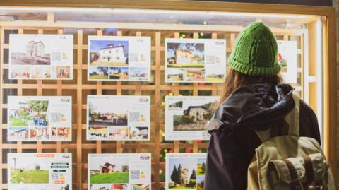 Woman wearing a green beanie hat looks at house prices in a store window