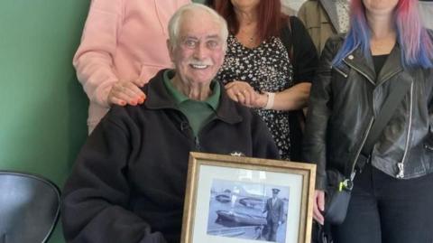 Stuart MacDonald smiling at the camera wearing a black work fleece and a green shirt, and holding a frame with an old black-and-white photo of him. A group of women, partially out of frame, stand behind him, with one resting a hand on his shoulder.