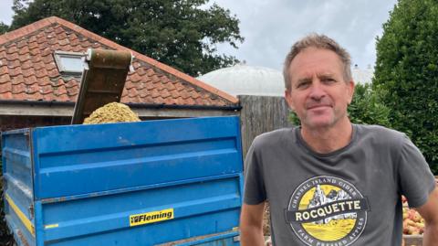 A man with grey hair wearing a grey and yellow t-shirt is standing in front of crates of red and green apples. There is a building in the background, and a large green bush.