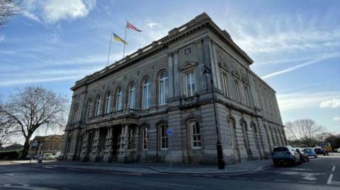 A grand building with long symmetrical windows going across the top and pillars out the front. Two flags are on top of it and several cars are parked around it.