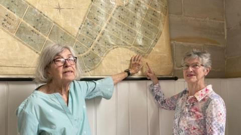 A woman in a light aqua shirt, wearing glasses, standing next to a woman who is wearing a multi-coloured shirt. They are pointing at a map reflecting the Tunbridge Wells Cemetery. 