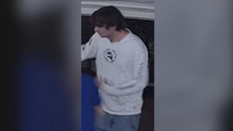 CCTV image of a young man with long dark hair wearing a white top wanted in connection with arson at Tesco in Hove