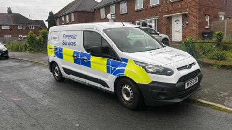 A police forensic van is pictured parked on a street outside residential homes. It is a white van with a yellow-and- blue chequered square pattern on its side. The words, "Forensic Services" are also written on its side and its bonnet.