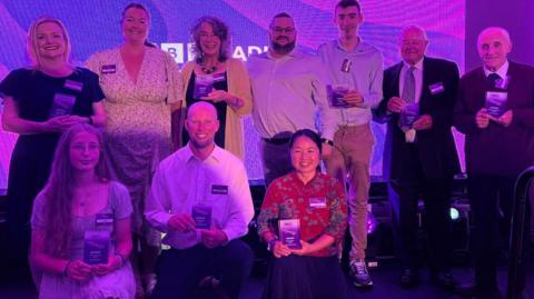 The winners photographed in a group at the ceremony, all smiling and smartly dressed holding their glass awards. Three of them are kneeling in front of a row of winners. They are standing in front of a screen which is purple and has the white BBC Radio Cornwall logo on it.
