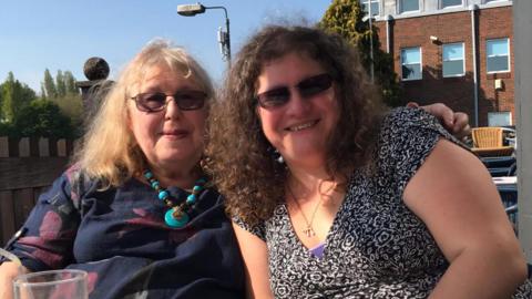 A woman (left) with blond hair, wearing a dark blue flowery top and turquoise necklace has her arm around her daughter (right) who wears a black and white top and has brown, curly hair. 