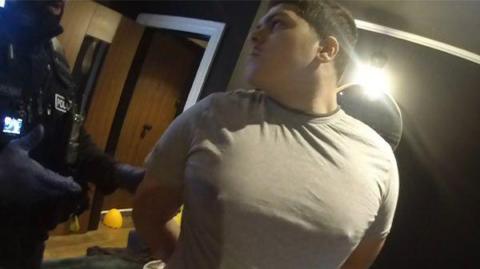 A still image from bodycam footage of Dearnie Jones being arrested. He is wearing a grey t shirt and has his hands behind his back.