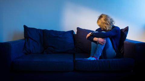 Posed by model - a woman is sitting on a sofa holding her knees under her chin with her head bowed.