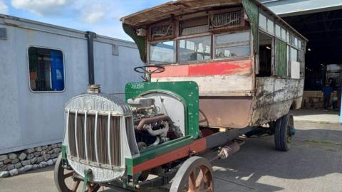 A photo of the old 1920s Eastern Counties bus that was made in Ipswich. It shows a singer decker bus with its engine at the front on show. The paint of the bus has been scratched off over time.