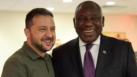 Presidents Volodymyr Zelensky and Cyril Ramaphosa are seen smiling as they pose for a picture. Zelensky is wearing a military green golf shirt while Ramaphosa dons a black suit, paired with a white shirt and purple tie.