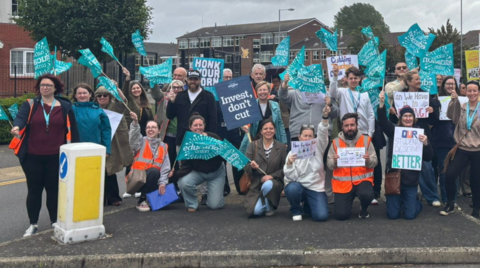 A group of dozens of people are at a road junction, with many holding light blue banners. Some are kneeling down and others are standing up, with buildings and trees in the background.