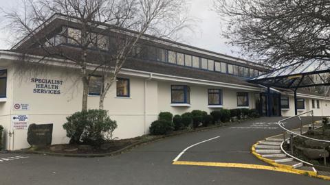A white building with blue framed windows, and a sign that reads 'specialist health services on it' a driveway leads up to an entrance.