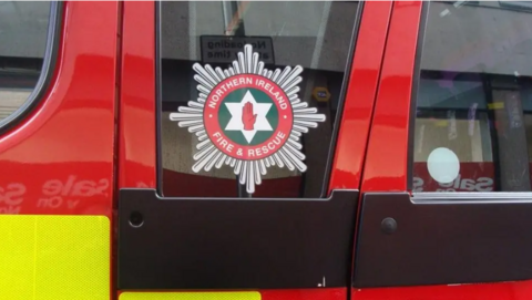 A red, yellow and black fire engine. The Northern Ireland Fire and Rescue Service (NIFRS) emblem is visible on a window of the vehicle.