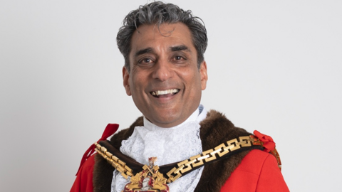 A man with dark and some grey hair is smiling at the camera. He is wearing mayoral clothing, including a mayoral chain, and standing in front of a plain white background.