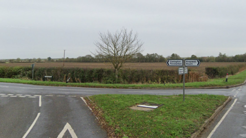 A three-way junction with a triangle patch of grass in the middle. In the background there is a brown field, sectioned off by a hedge and tree.