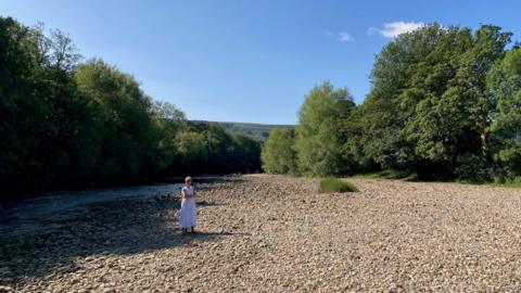 A woman in a white dress standing among thousands of pebbles on a dry river bed with a stream of water running beside her.