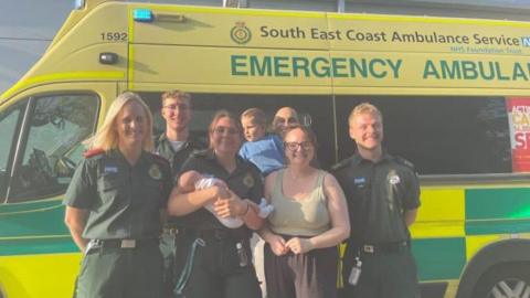 Paramedics stand with the family in front of an ambulance. One paramedic is holding baby Otis, while the father, Joe, is holding toddler Theo.