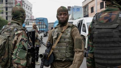 Armed M23 fighters stand in a street wearing military fatigues. Two are stood with their backs towards the camera, while one faces the opposite direction.