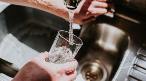 A person fills a clear glass with running water from a kitchen tap. One hand holds the glass, the other adjusts the tap. The sink is metallic with visible water splashes.
