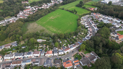 An aerial view of rows of houses on opposite sides of a main road, surrounded by fields and trees in Jersey.