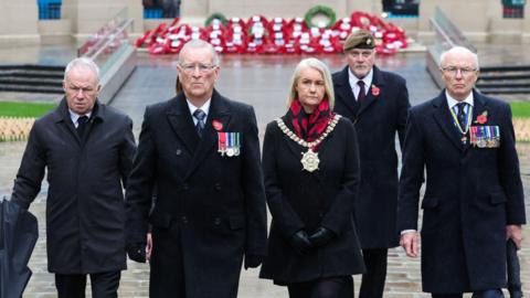 Tracy Kelly is wearing the mayoral chain of Belfast. She has shoulder length blonde hair and is dressed in black with a red and black scarf. One either side of her are two men dressed in black overcoats decorated with poppies and military medals. There are two other men also dressed in black, one of whom is wearing a military beret. In the background there are several rows of poppy wreathes.