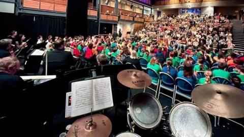 A set of drums at the front of the stage. There is people sat in different coloured jumpers on chairs facing an audience of people looking back at them.