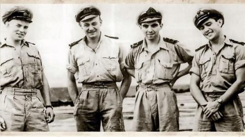A black and white photograph of four young men in army uniform. They have their sleeves rolled up and are wearing military caps. They are standing around looking relaxed and are smiling.