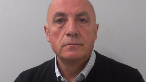 A bald man wearing a blue shirt with a black jumper on top. It is a police mugshot.