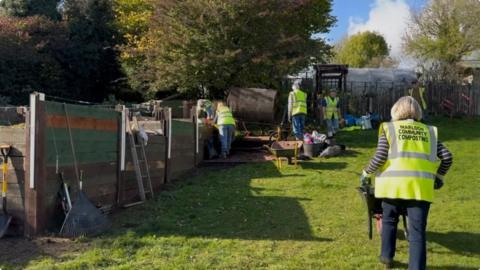 Five volunteers, dressed in jeans and luminous green, high-vis tabards, doing various jobs on site.  One is pushing a wheelbarrow, another two are working by a soil tumbling machine, two others are checking the state of the compost.
