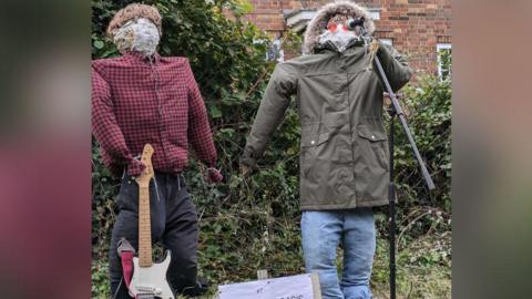 Liam Gallagher's signature parka and a pair of sunglasses is worn by one of the scarecrows, while the other is holding a guitar and wearing a checked shirt and a wig to replicate Noel Gallagher's hairstyle.