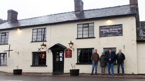 Three men and a woman are standing outside a pub that is painted white and has black windows. 