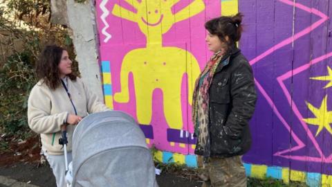 Two women, one pushing a pram, stand next to a bright purple and yellow mural
