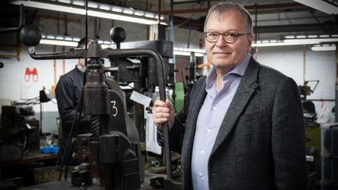 A man wearing glasses, a grey blazer and a light blue shirt stands beside a heavy iron fly press in an industrial workshop. Behind him, strip lighting illuminates a cluttered workspace filled with machinery, tools and equipment. A second worker can be seen in the background on the left of the frame.