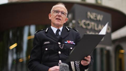 A balding middle aged man wearing police uniform stands outside New Scotland Yard police headquarters. He is reading a statement from a clipboard which he is holding in his left hand.